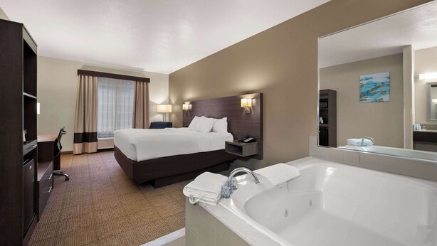 A hotel room with a large bed, a desk, a chair, a desk lamp, a nightstand, a telephone, a bathroom with a bathtub, a sink, and a mirror. - Comfort Inn & Suites Fenton (Fenton)