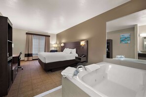 A hotel room with a large bed, a desk, a chair, a desk lamp, a nightstand, a telephone, a bathroom with a bathtub, a sink, and a mirror. - Comfort Inn & Suites Fenton (Fenton)
