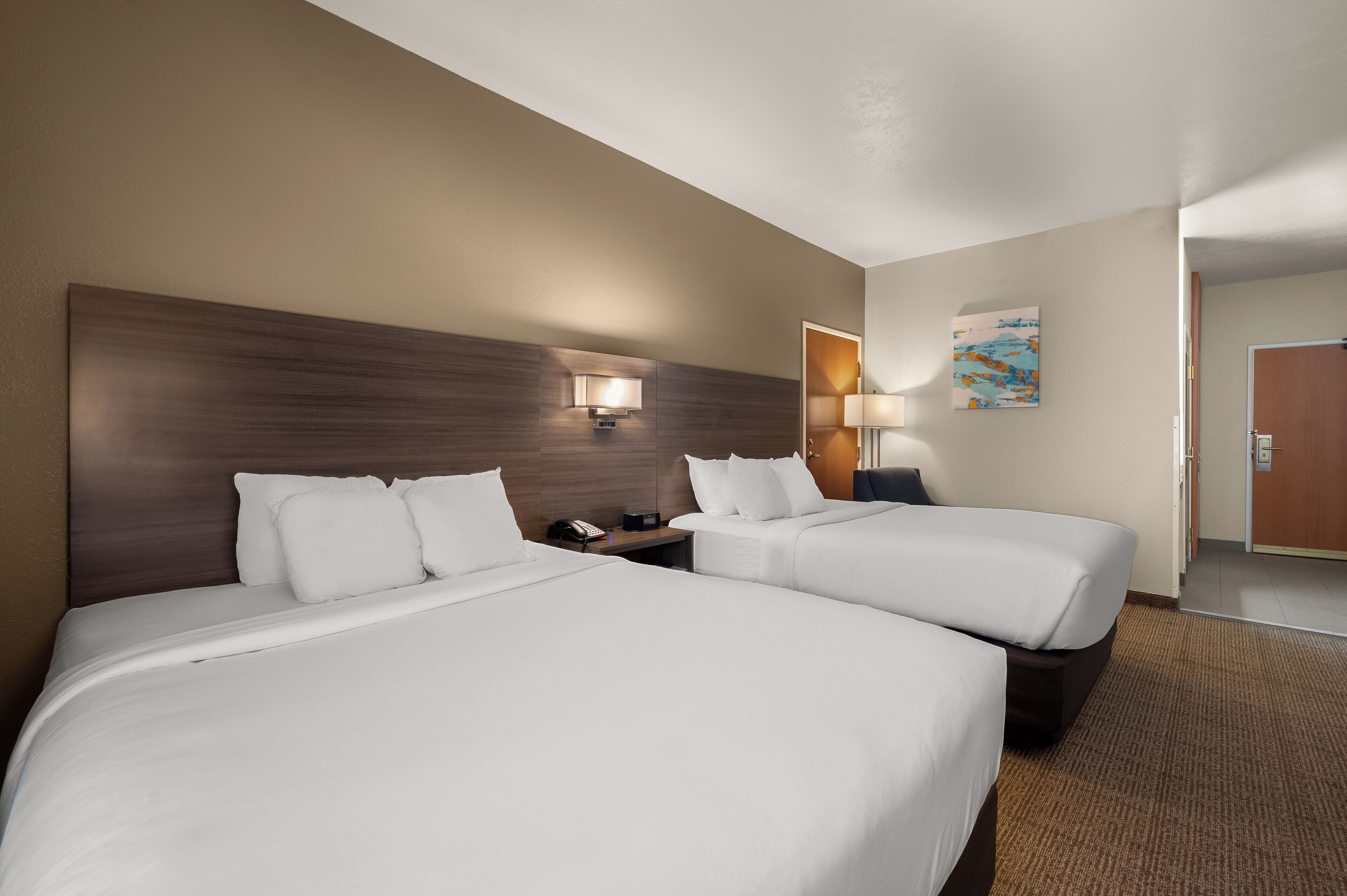 A hotel room with two beds, a wooden headboard, a nightstand, a lamp, and a painting on the wall.