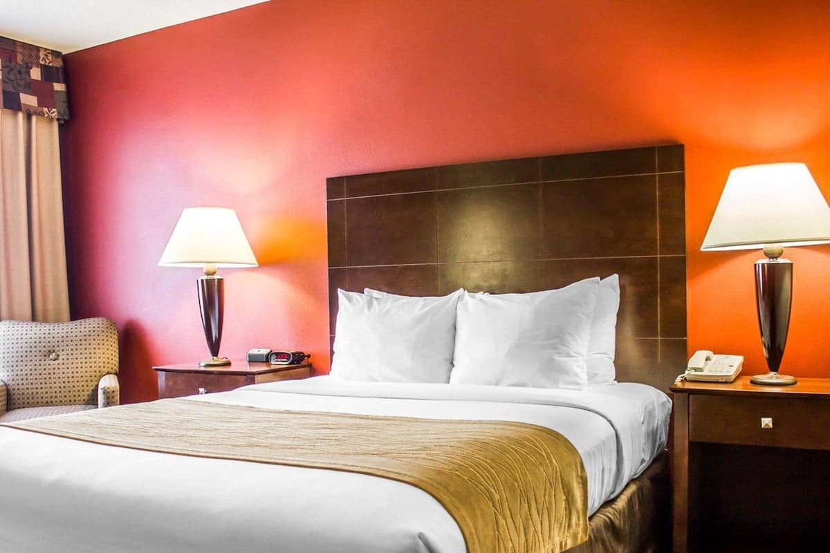 A hotel room with a large bed, two bedside lamps, a nightstand, a chair, and a red wall.