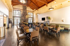 A spacious restaurant with high ceilings and exposed beams, featuring multiple dining tables and chairs, a stone accent wall, and a large window allowing natural light.