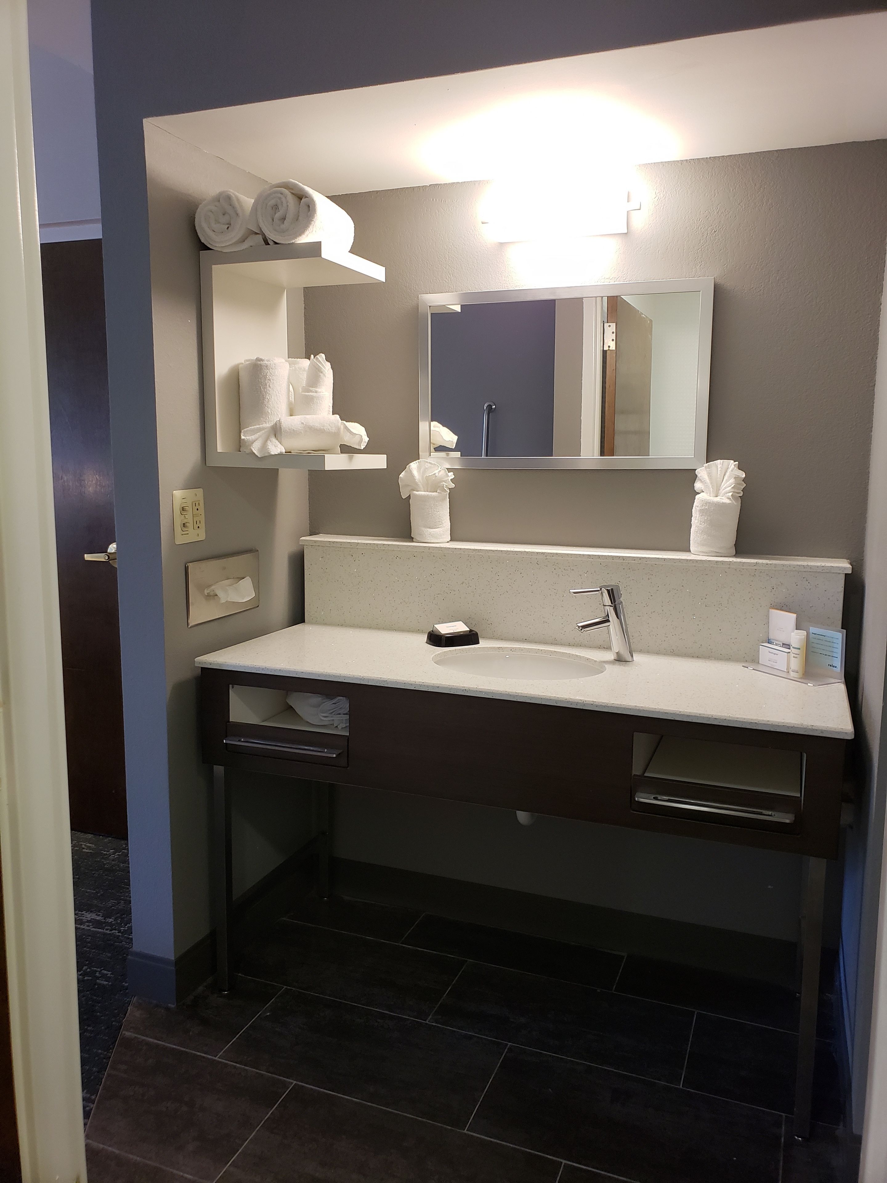 Un baño moderno con un lavabo blanco, encimera oscura y un espejo grande sobre él. Hay toallas y un dispensador de jabón en la encimera.