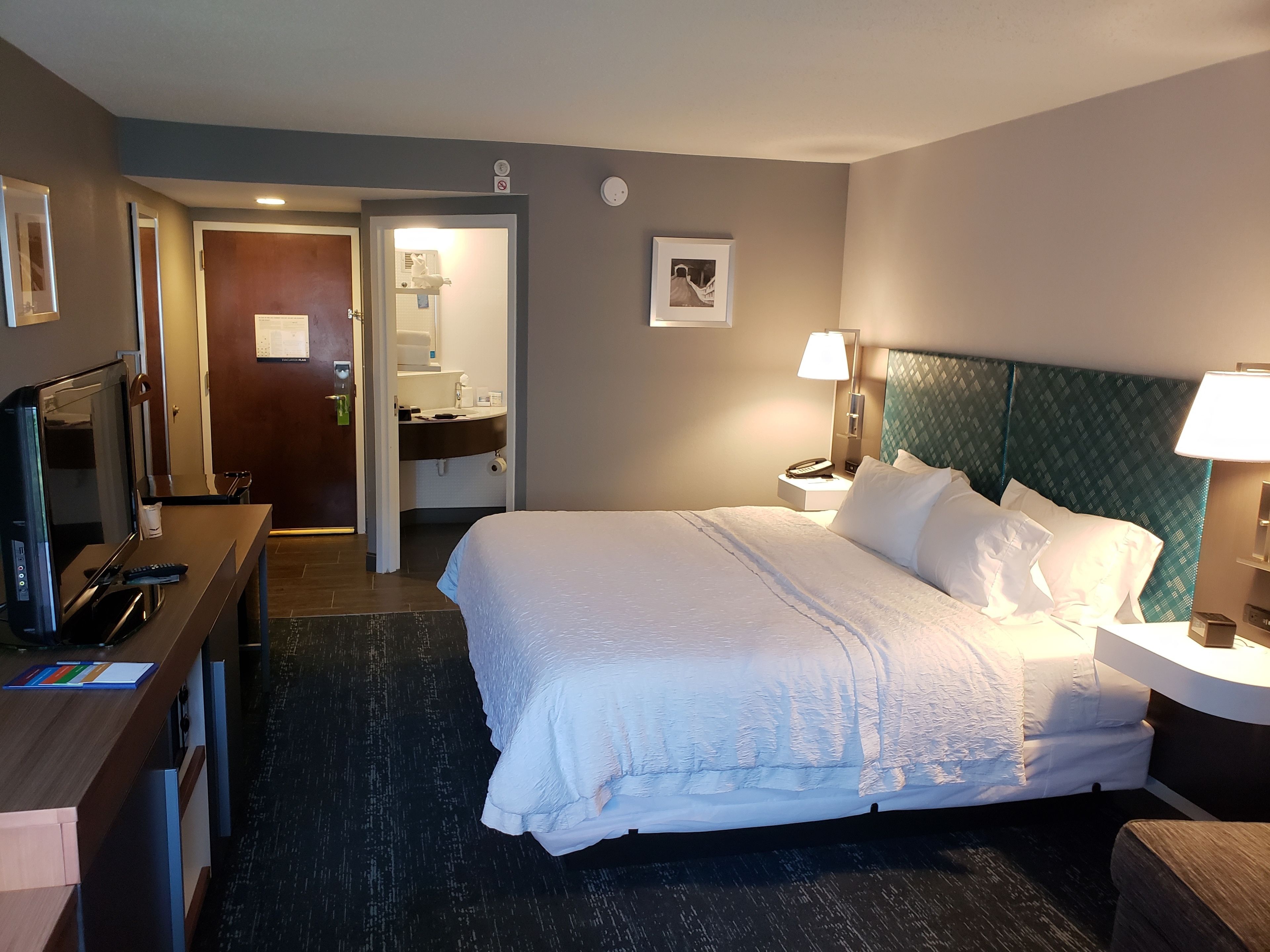 A hotel room with a bed, bedside table, lamp, and a television on a desk.