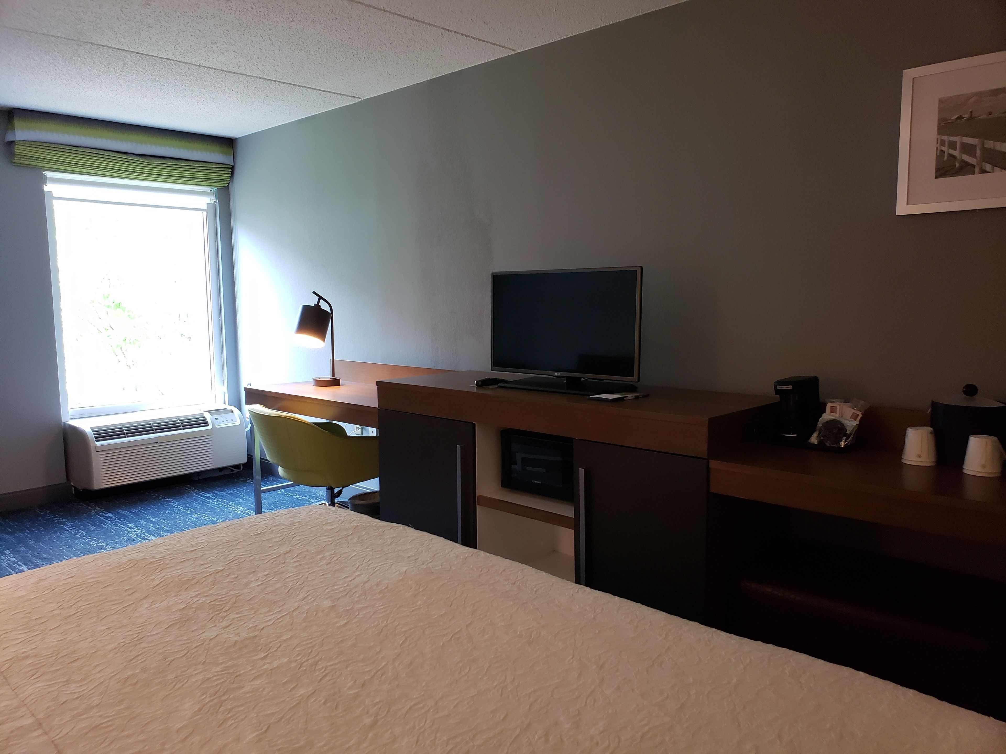 A hotel room with a bed, a desk with a computer, a chair, a TV, and a window with blinds.