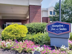 A Hampton Inn & Suites sign with a garden of colorful flowers and a brick building in the background.