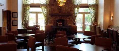 A restaurant with a stone fireplace, wooden ceiling beams, and a hanging light fixture. Tables and chairs are arranged for dining.