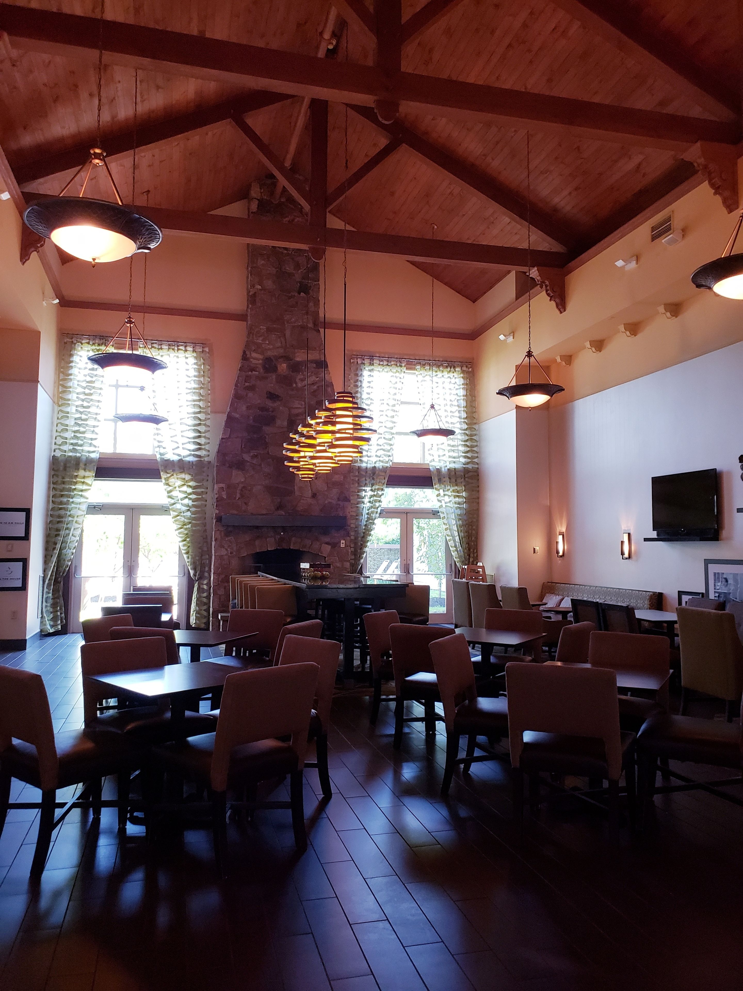 A restaurant with wooden beams, hanging pendant lights, stone walls, and a fireplace. Tables and chairs are arranged for dining.