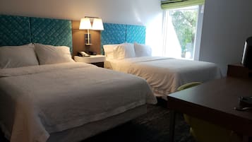 Ein Hotelzimmer mit zwei Betten, einem Schreibtisch, einem Stuhl, einer Lampe und einem Fenster mit Blick auf Bäume.