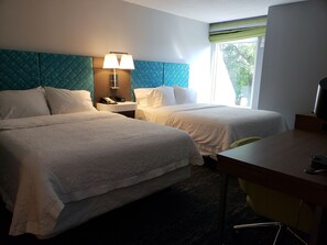 A hotel room with two beds, a desk, a chair, a lamp, and a window with a view of trees.