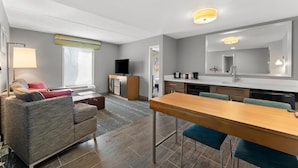 A modern living room with a sofa, a dining table, and a kitchen area with a TV and appliances. - Hampton Inn & Suites Newtown (Morrisville)