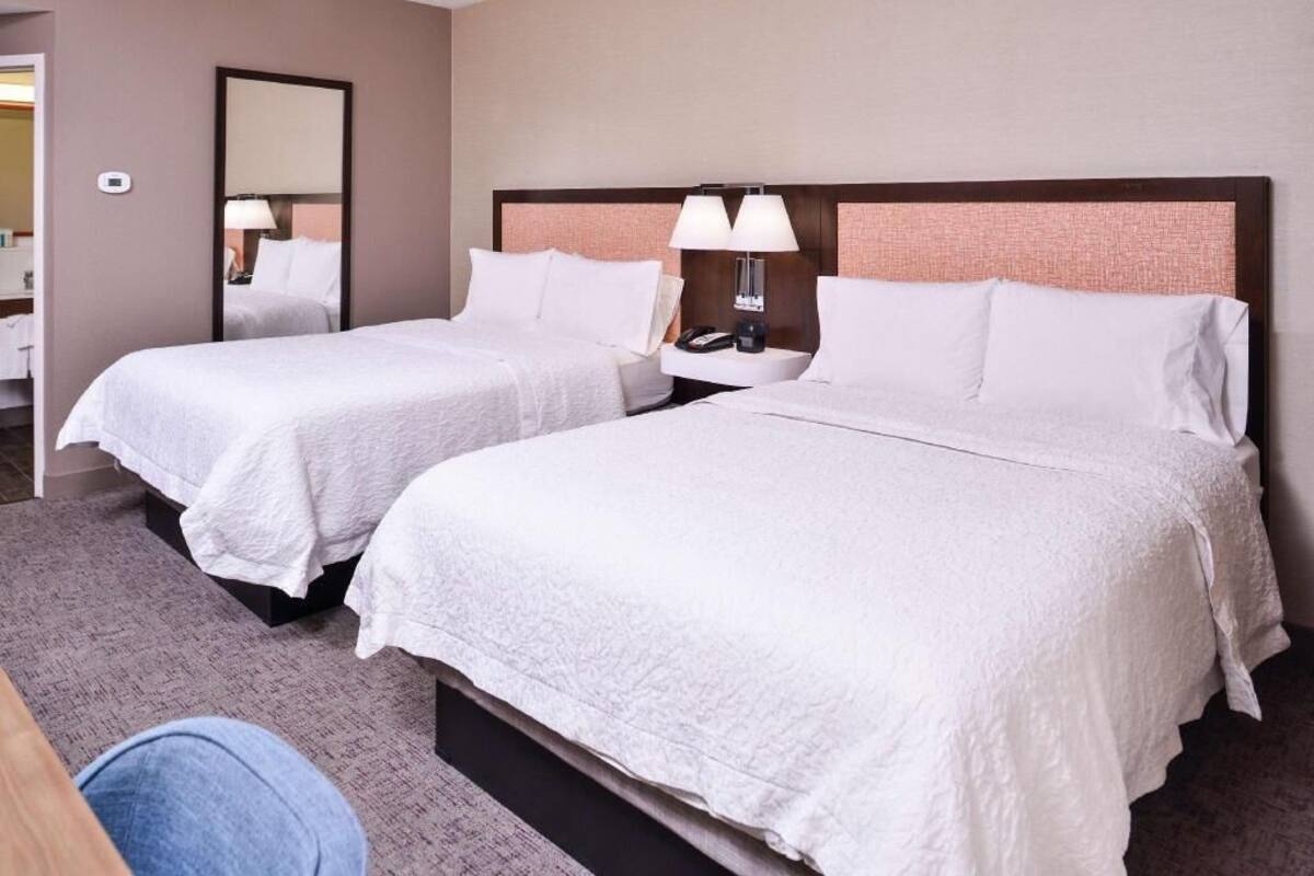 A hotel room with two beds, a nightstand with a lamp, a mirror, and a carpeted floor.
