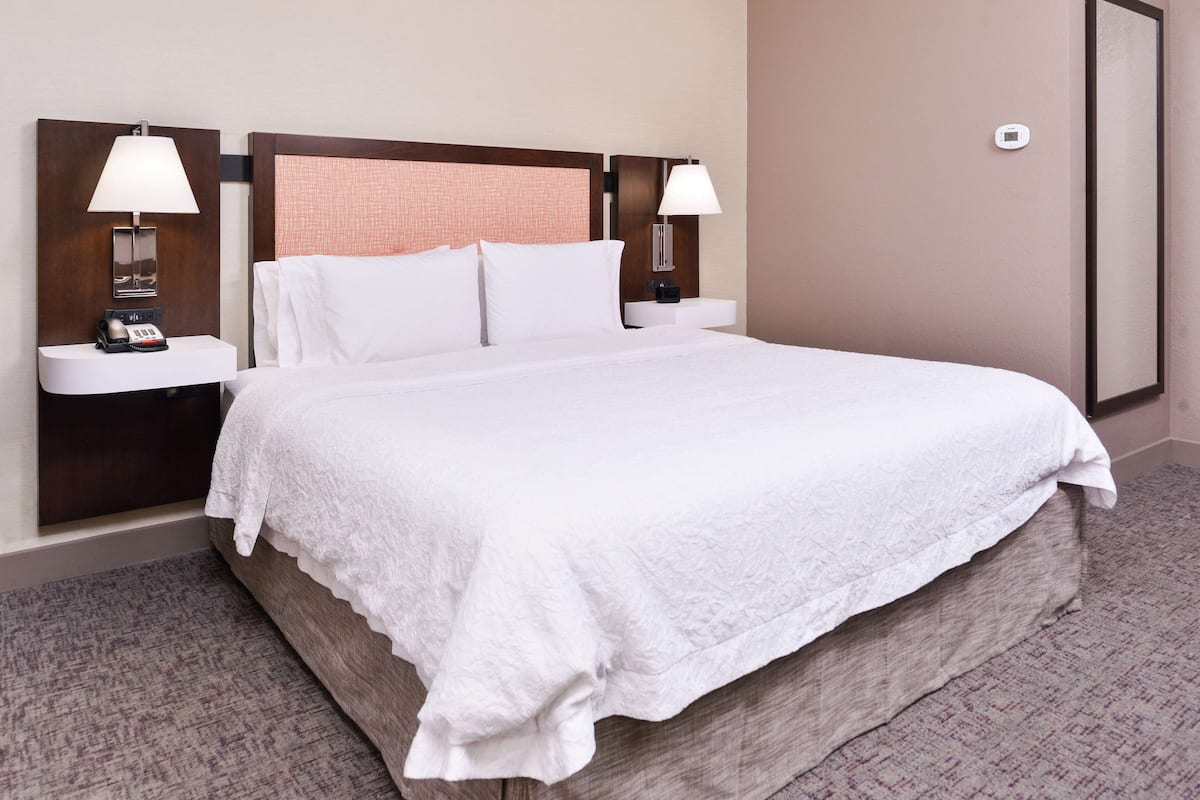 A hotel room with a large bed, two bedside tables with lamps, a thermostat, and a framed picture on the wall.