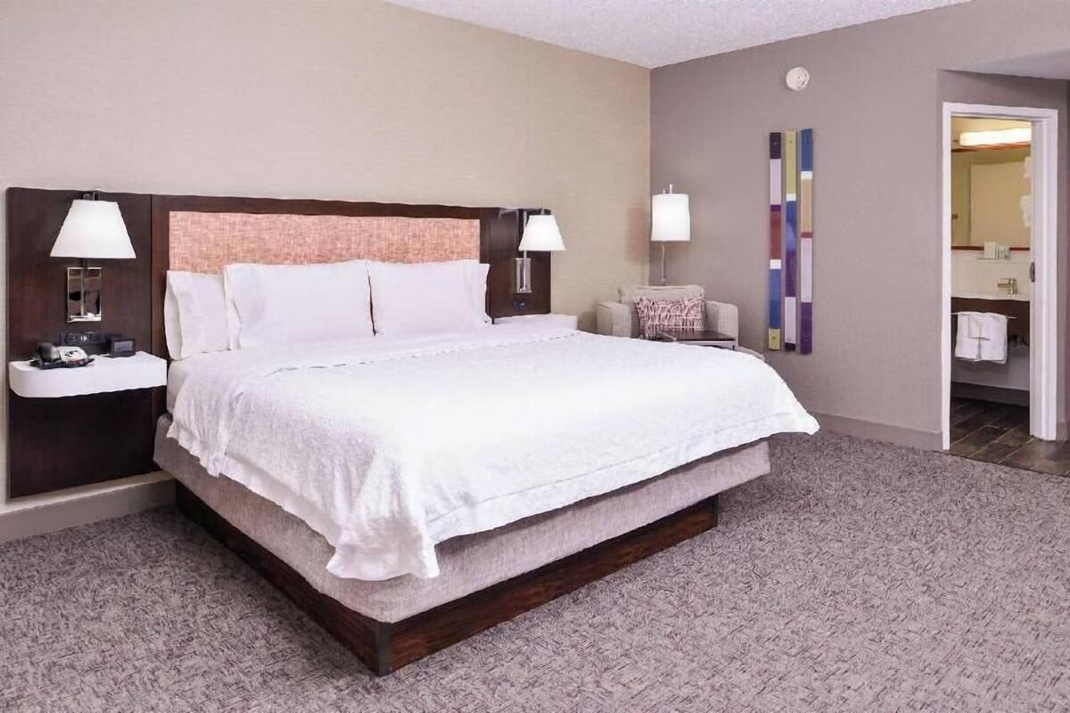 A hotel room with a large bed, two bedside tables with lamps, a small seating area, and a bathroom visible through an open door.