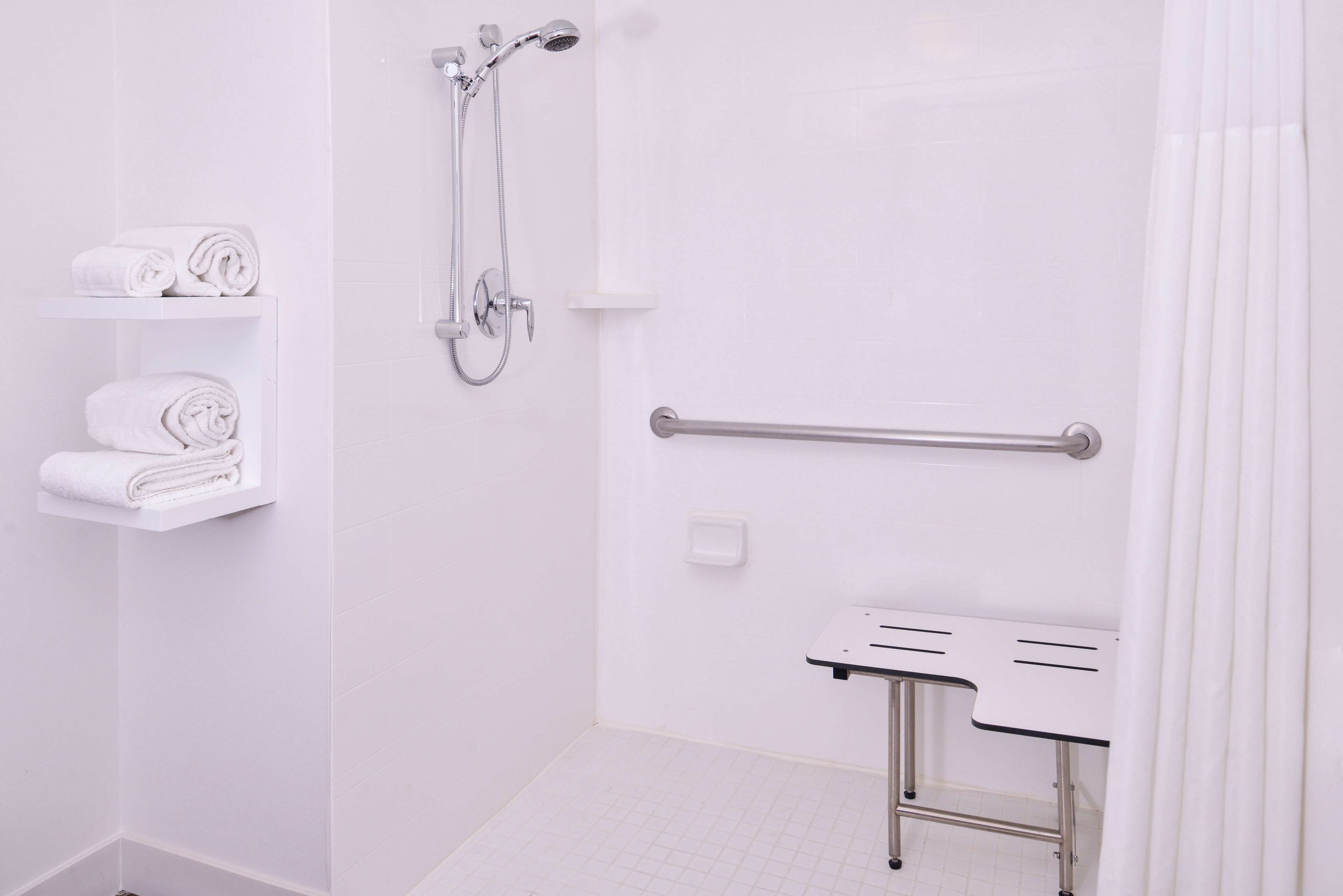 A white bathroom with a shower, a toilet with a grab bar, and shelves with folded towels.