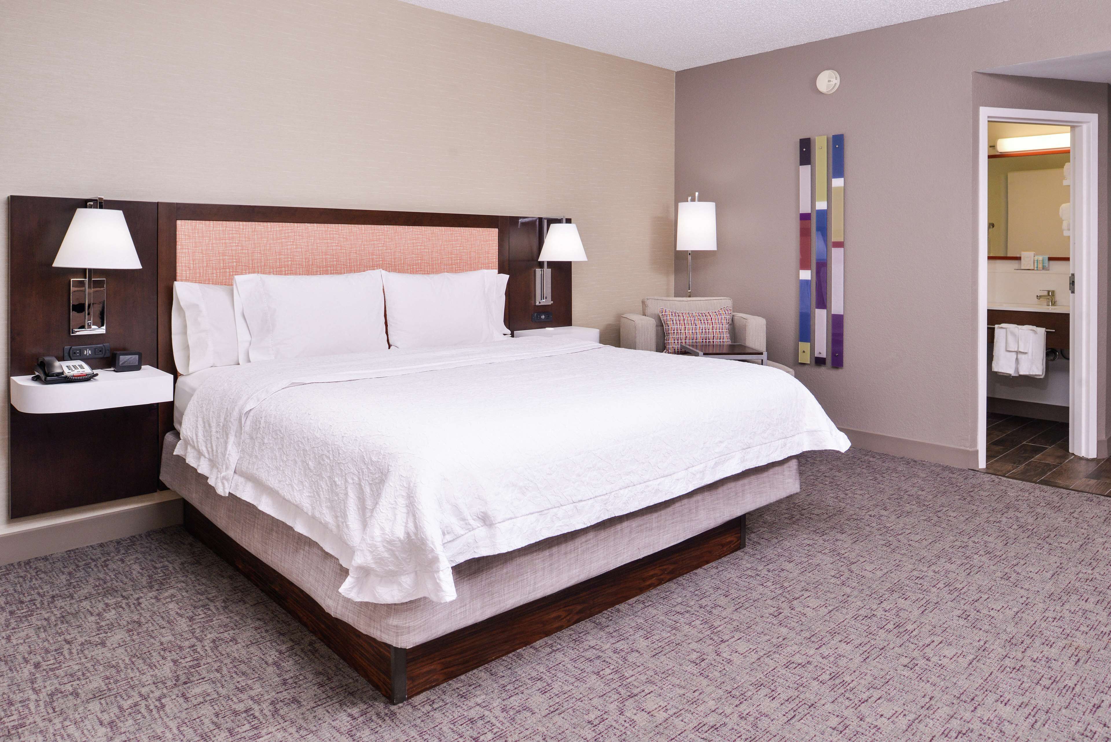 A hotel room with a large bed, two bedside tables with lamps, a small armchair, and a bathroom visible through an open door.