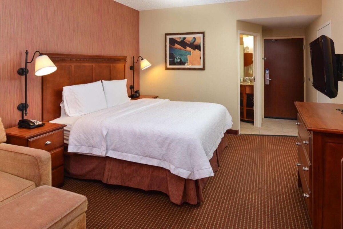 A hotel room with a bed, bedside lamps, a wooden headboard, a painting on the wall, a television, and a desk with a telephone.