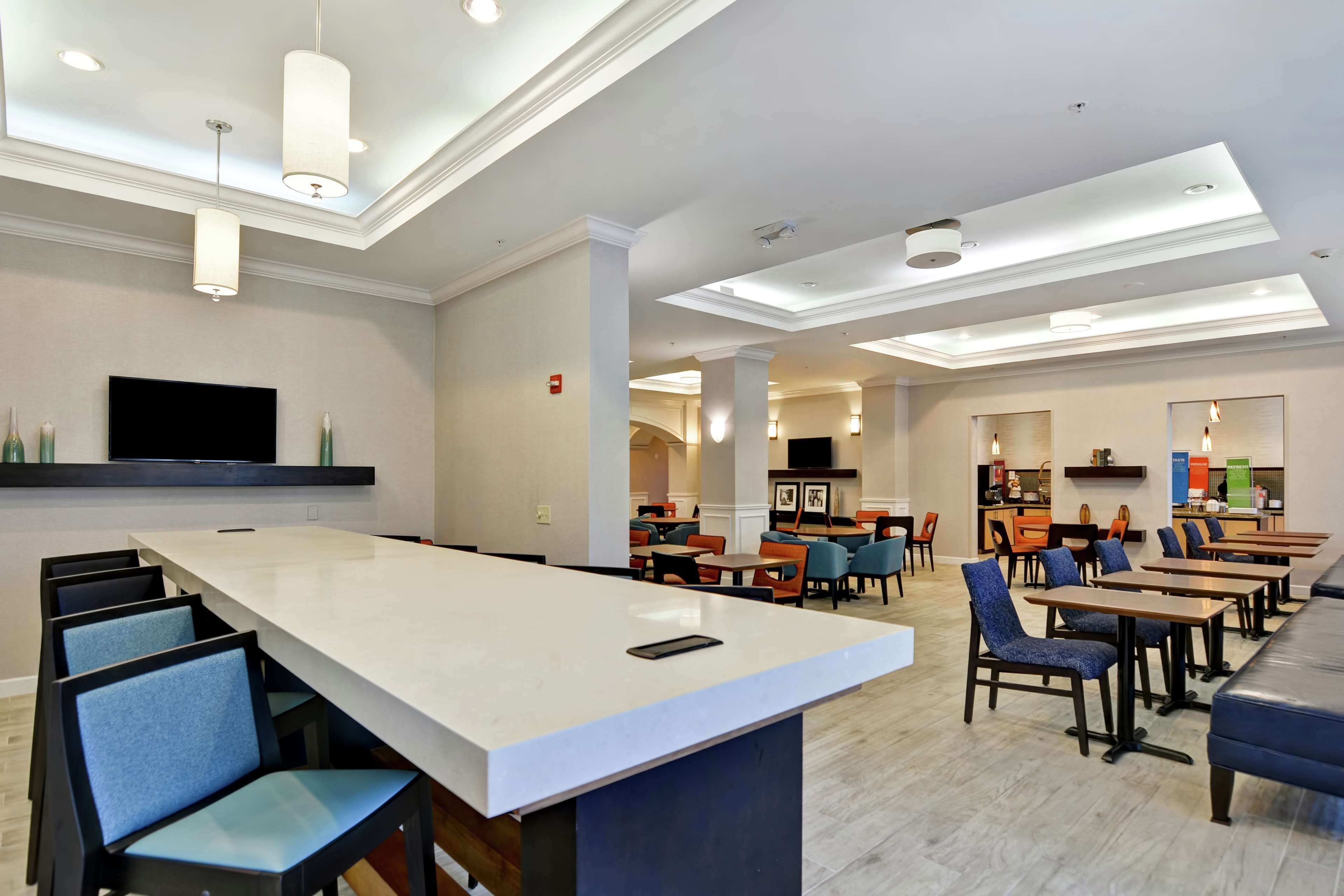 Un hall d’hôtel moderne doté d’un bar central, de différents espaces aménagés pour s’asseoir et d’un téléviseur à écran plat fixé au mur.