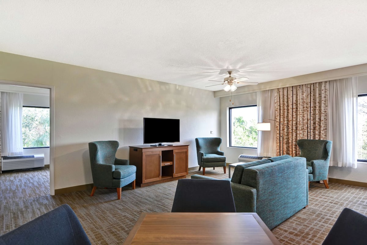 Habitación de hotel con televisor, dos sillones, un sofá, una mesa de comedor y un ventilador de techo.