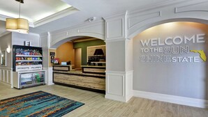 A hotel lobby with a reception desk, a 'Welcome to the Sun Shine State' sign, a vending machine, and a colorful rug. - Hampton Inn Palm Beach Gardens (Palm Beach Gardens)