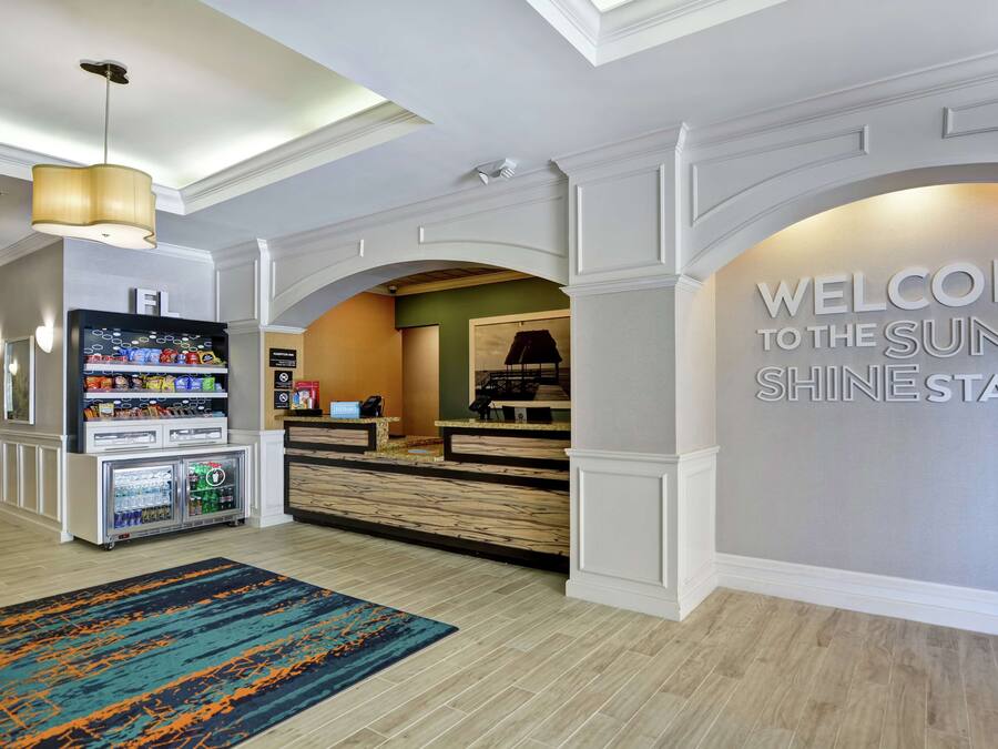 A hotel lobby with a reception desk, a 'Welcome to the Sun Shine State' sign, a vending machine, and a colorful rug.