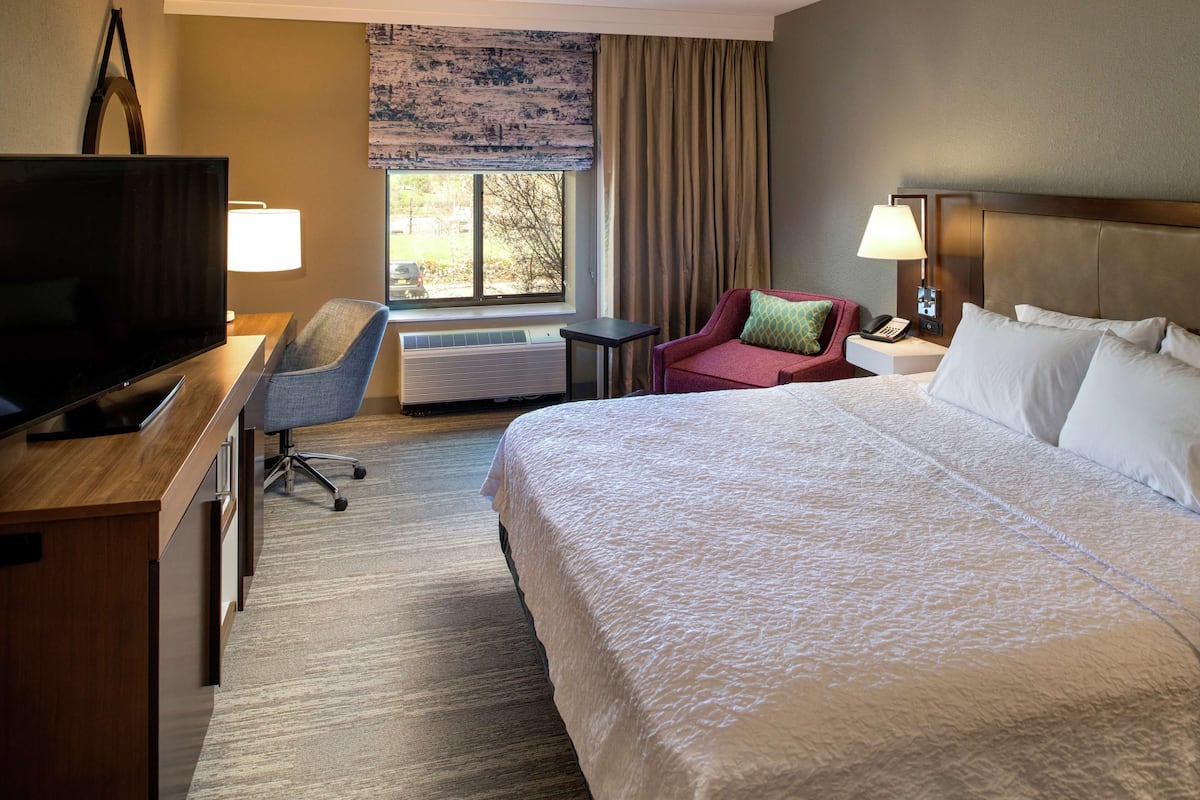 A hotel room with a large bed, a desk with a television, a chair, a window with curtains, and a nightstand with a lamp.