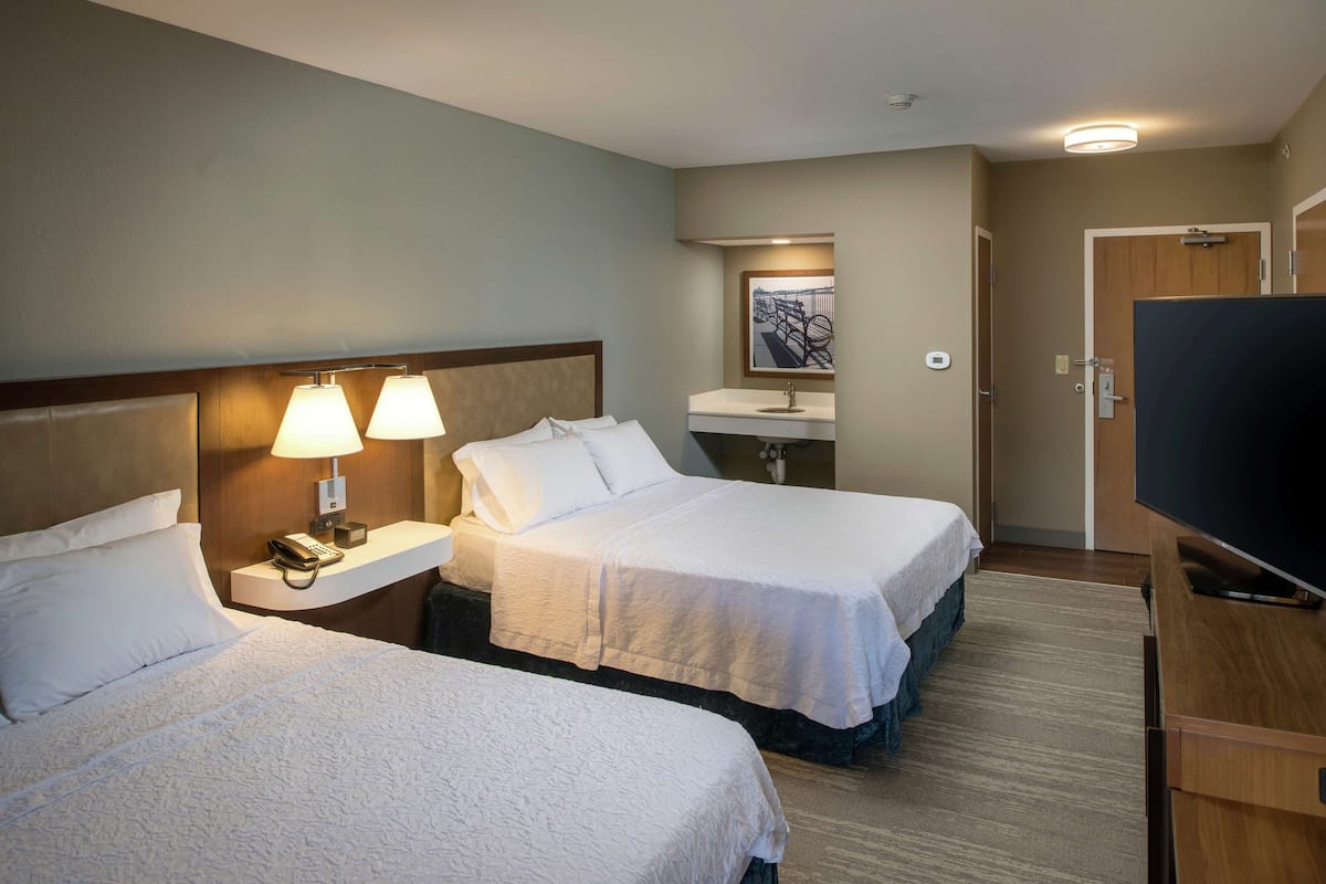 A hotel room with two beds, a desk with a lamp, a TV, a bathroom, and a painting on the wall.