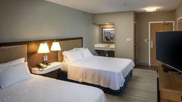 Et hotelværelse med to senge, et natbord med lampe, et fjernsyn og et badeværelse med vask og spejl.