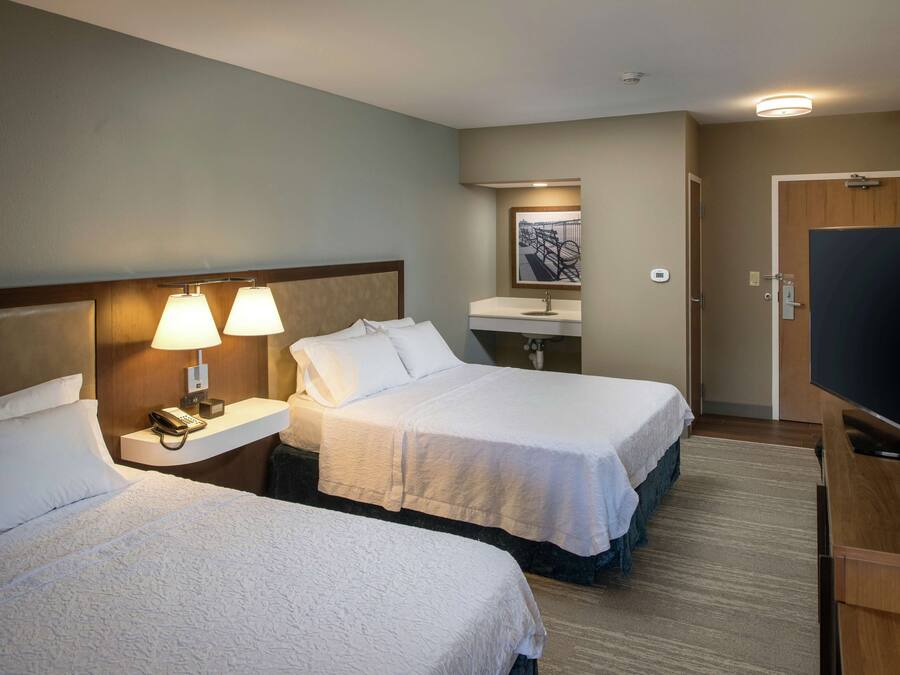 A hotel room with two beds, a bedside table with a lamp, a TV, and a bathroom with a sink and mirror.