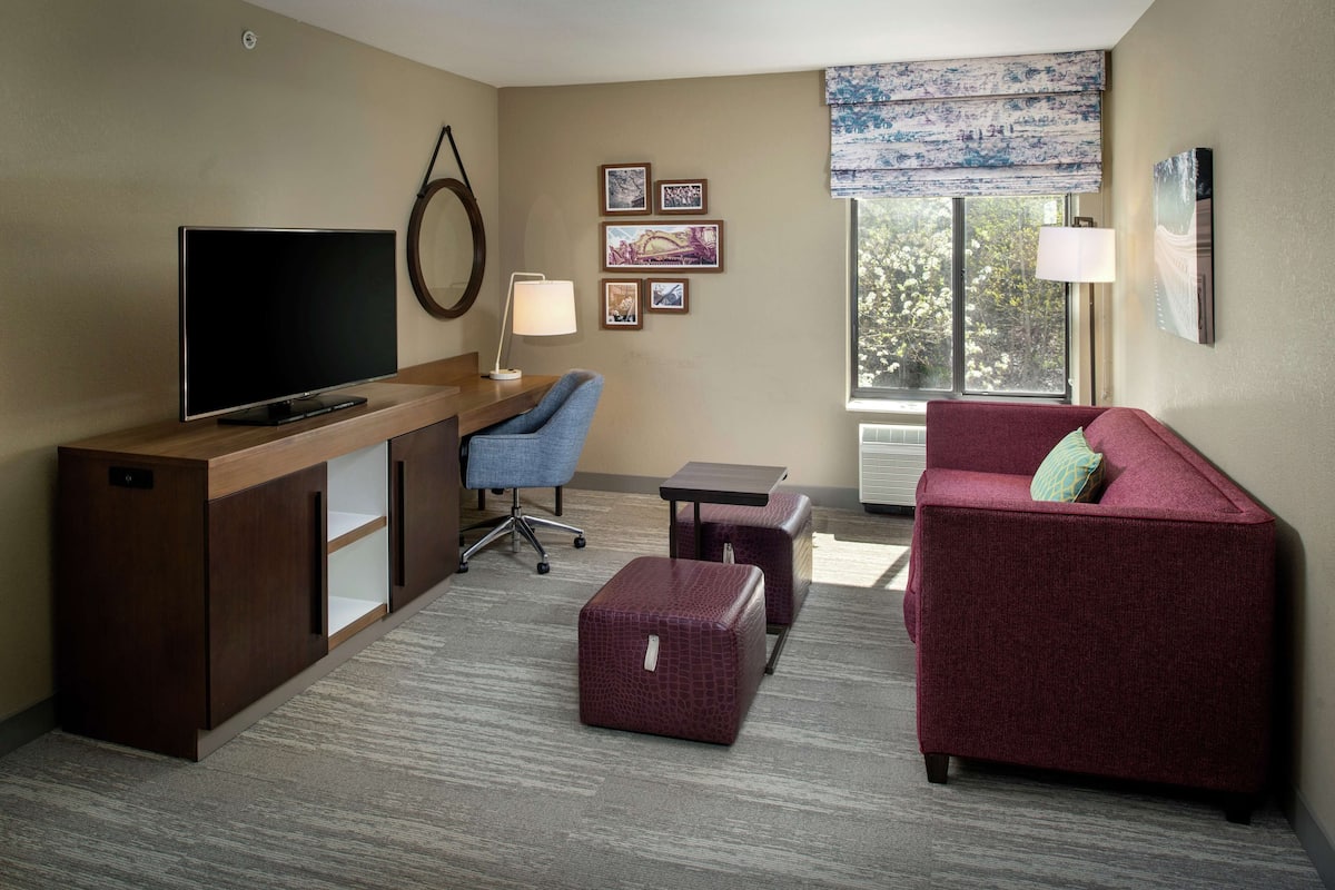 A hotel room with a wooden desk, a flat-screen TV, a blue office chair, a red sofa, a small table, and a window with a view of trees.