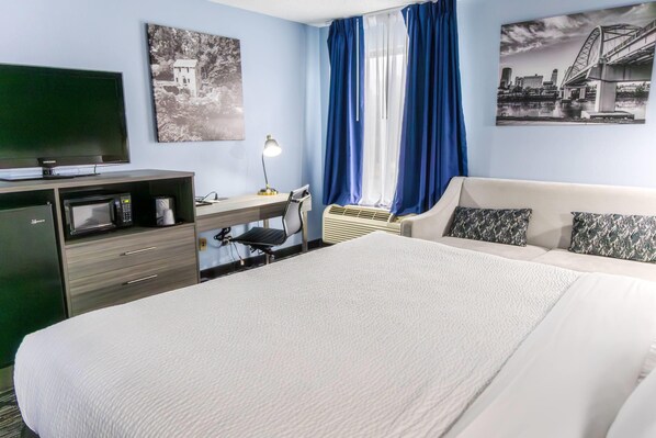 A hotel room with a bed, a desk with a computer, a television, a microwave, and a window with curtains.
