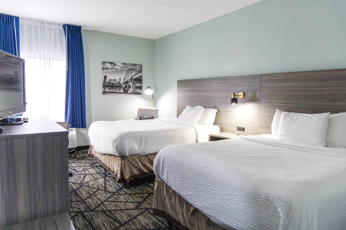 Une chambre d’hôtel avec deux lits, un bureau, une chaise, une lampe et un tableau représentant un paysage urbain accroché au mur.