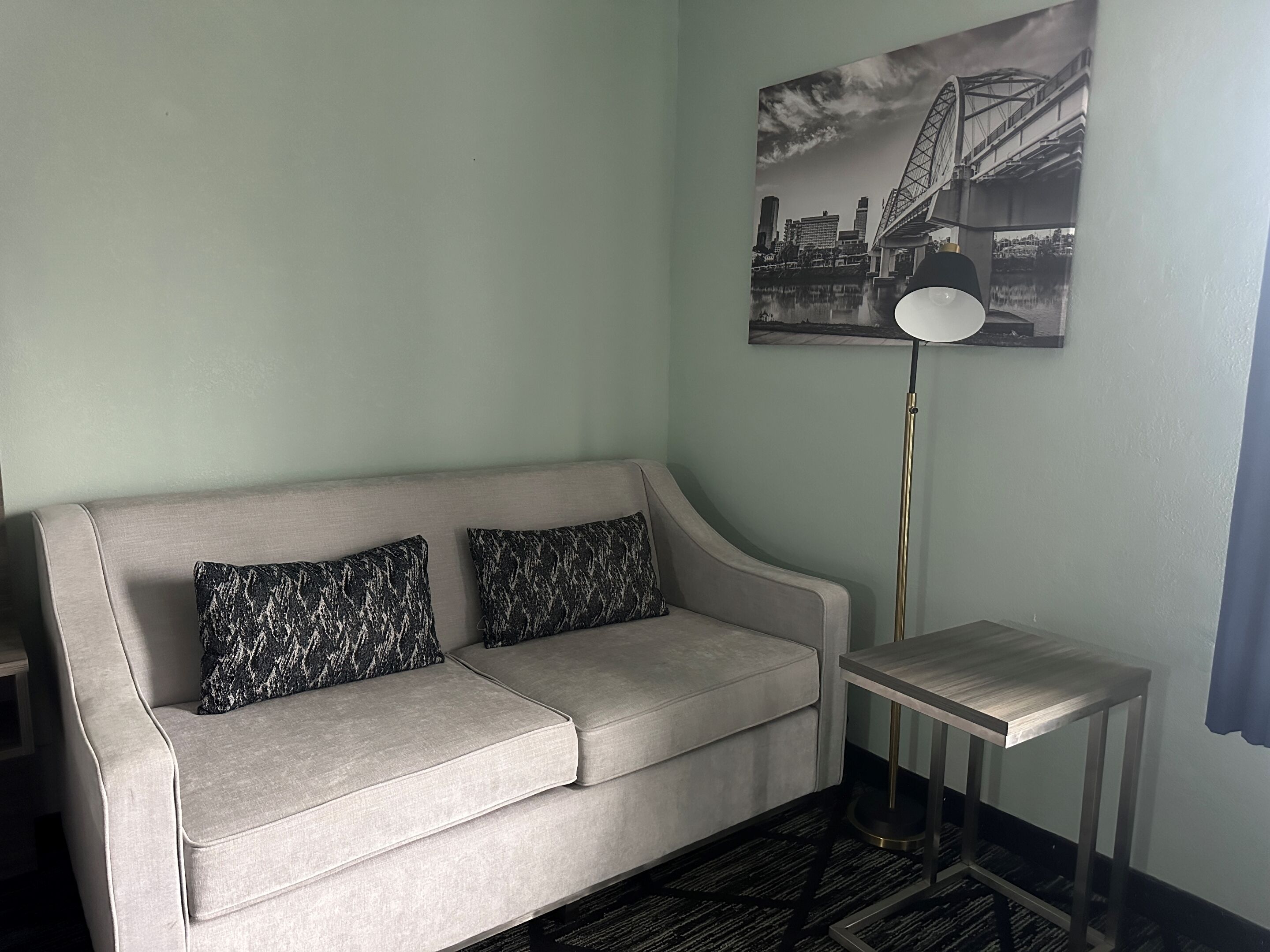 A room with a light grey sofa, two patterned pillows, a black and white cityscape painting, a tall floor lamp, and a small wooden side table.