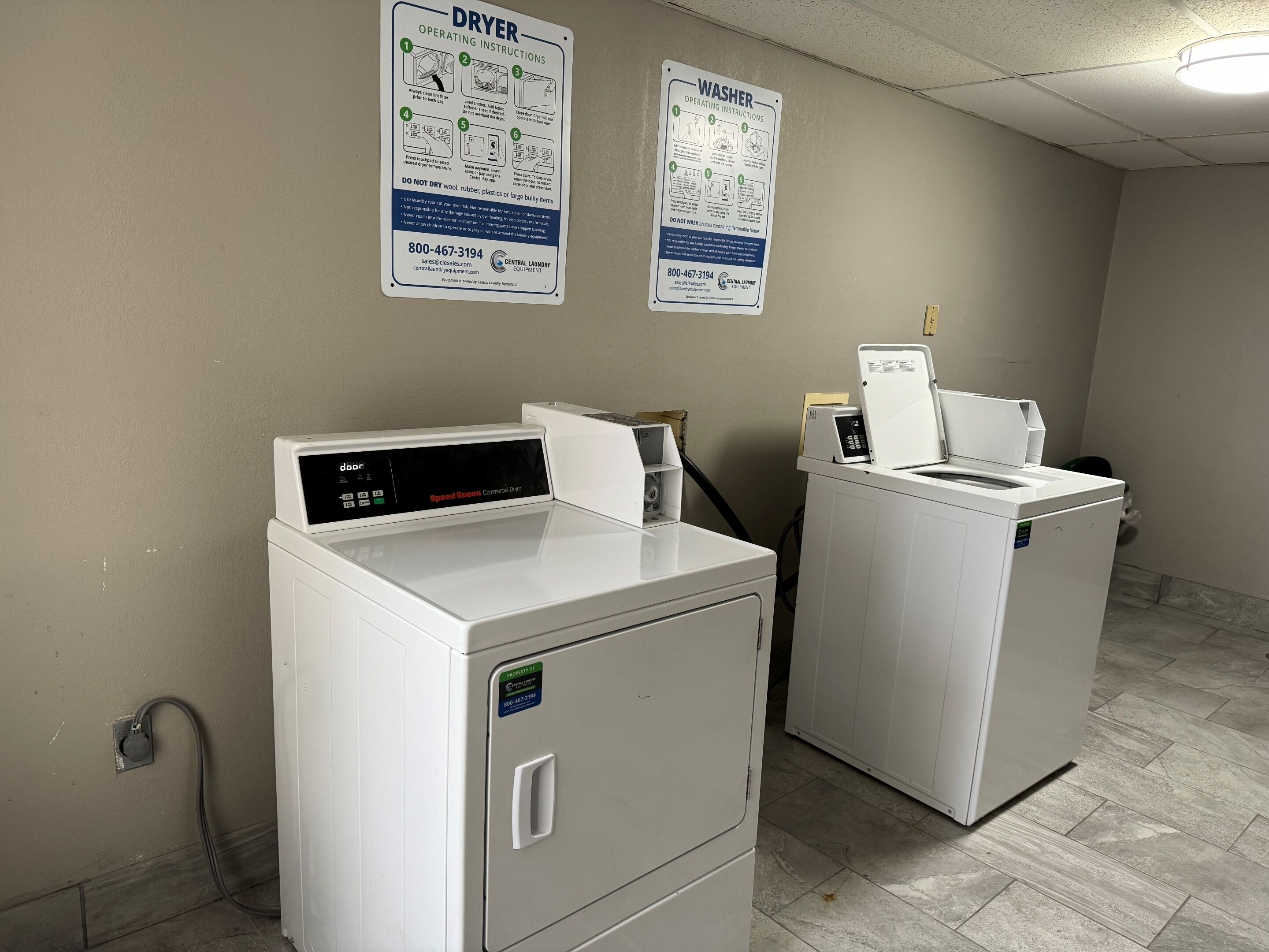 A laundry room with two front-loading washing machines, a dryer, and two instructional posters on the wall.