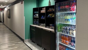 Un pasillo con una máquina expendedora llena de bebidas, un refrigerador pequeño y un expositor con aperitivos.