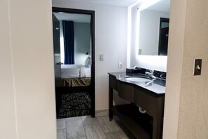 Una habitación de hotel con una cama, un baño con lavamanos y espejo, y una puerta que da a otra habitación.