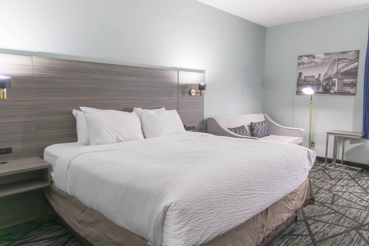 Une chambre d’hôtel avec un grand lit, une chaise, une table de chevet, une lampe et un tableau représentant un paysage urbain accroché au mur.