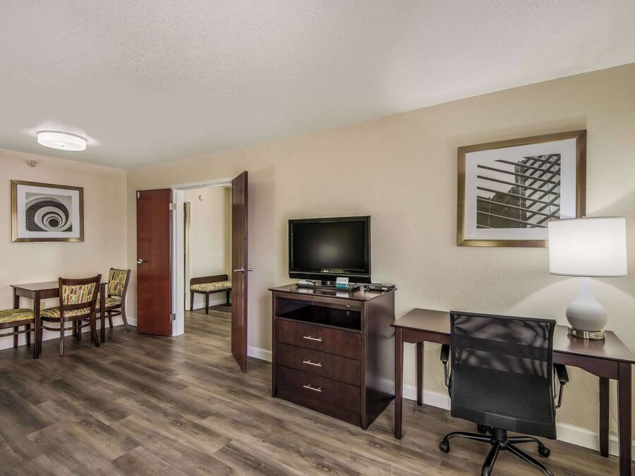 Quarto de hotel com escrivaninha de madeira, televisão, cadeira, luminária e área de refeições com cadeiras.