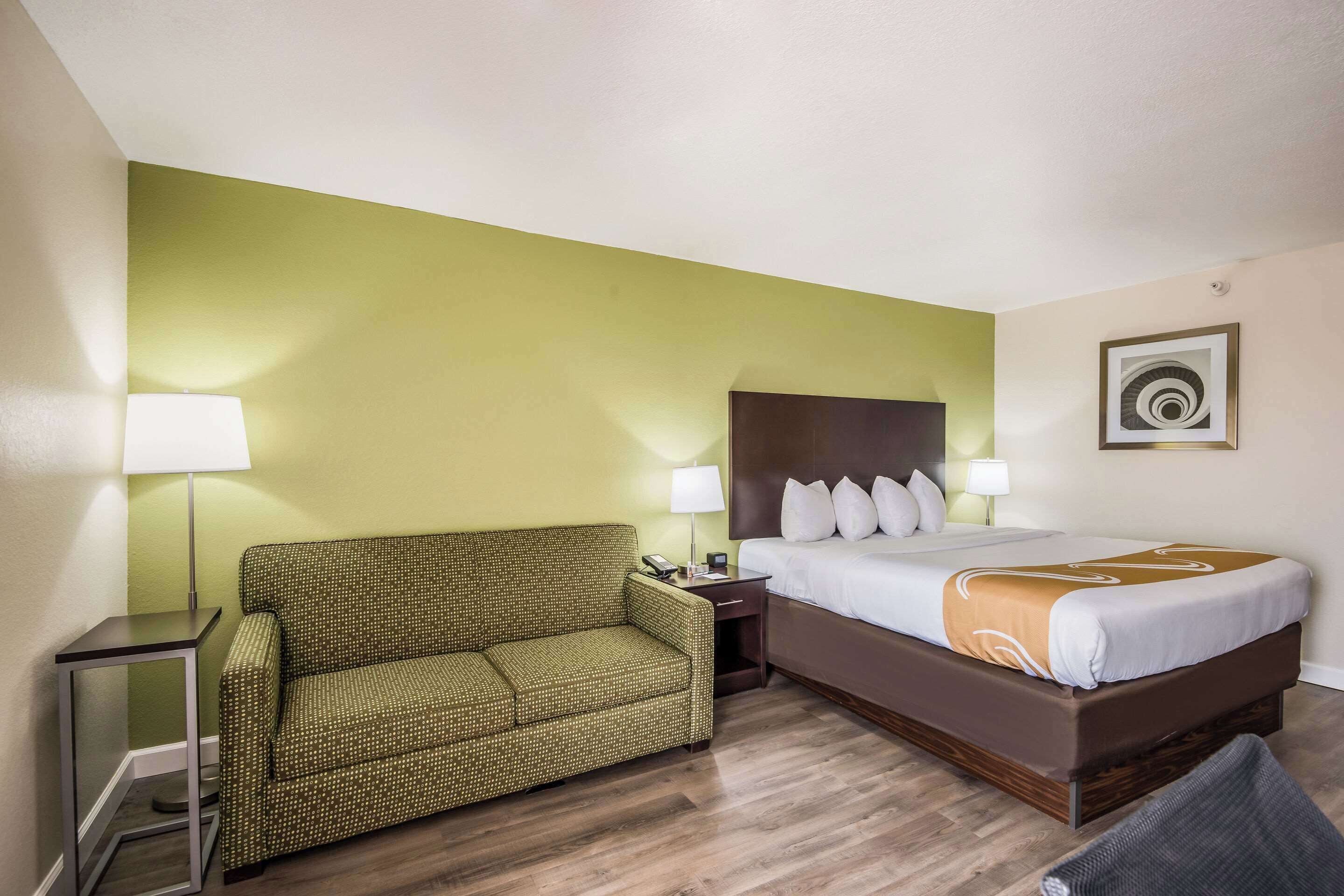 Una habitación de hotel con cama, un sofá verde estampado, una mesita de noche y una lámpara.