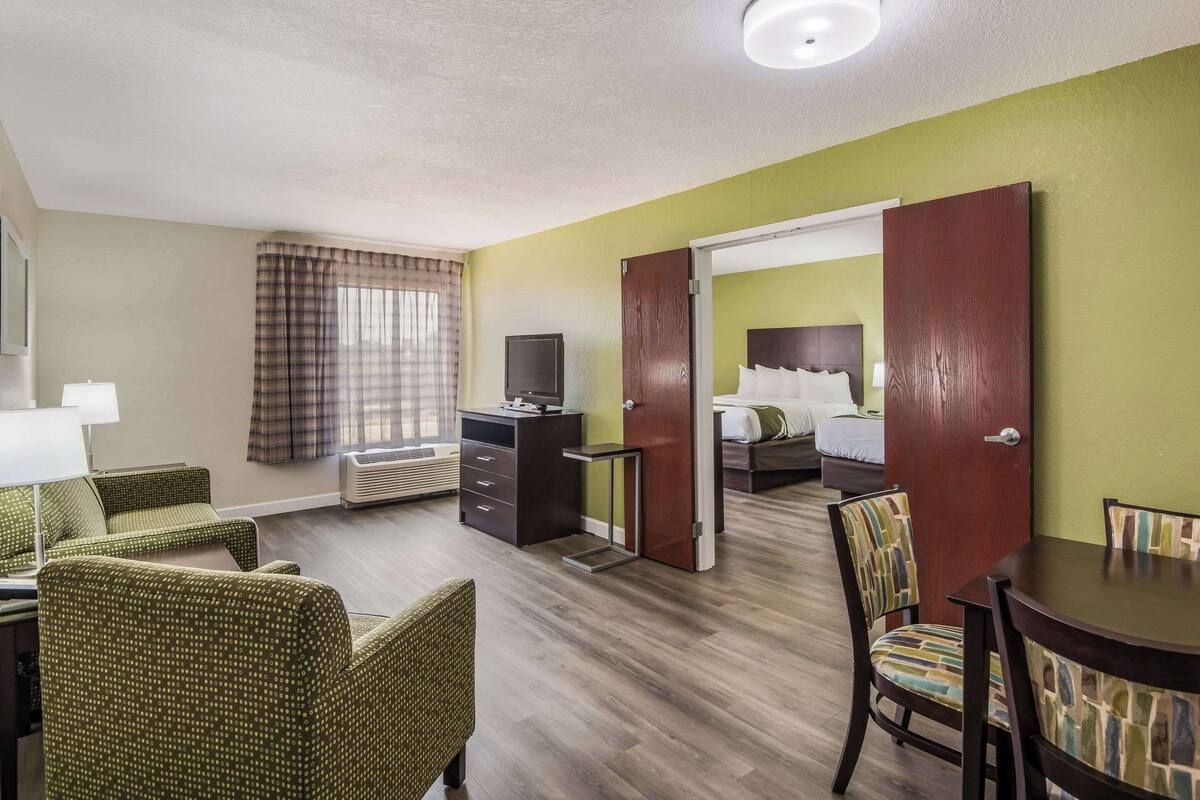 A hotel room with a bed, a desk with a chair, a television, and a dining area with chairs.