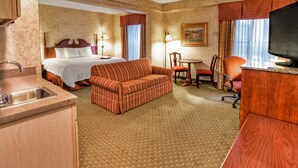 A hotel room with a bed, a sofa, a dining table with chairs, a television, and a kitchen area with a sink.