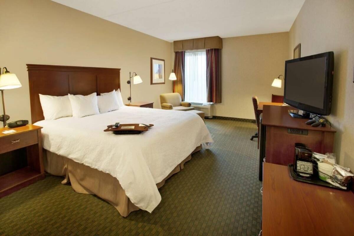 Una habitación de hotel con una cama grande, un escritorio con televisor de pantalla plana, una silla, una lámpara de escritorio y una ventana con cortinas.