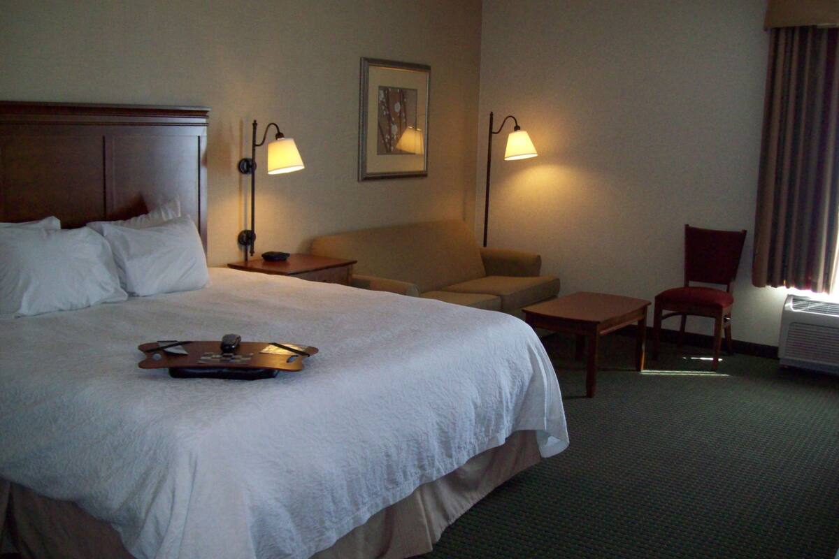 Quarto de hotel com uma cama grande, um sofá, uma poltrona, uma mesinha e duas luminárias fixadas na parede.