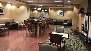 A dining area with wooden tables and chairs, a green cushioned bench, and pendant lighting. - Hampton Inn Sidney (Sidney)