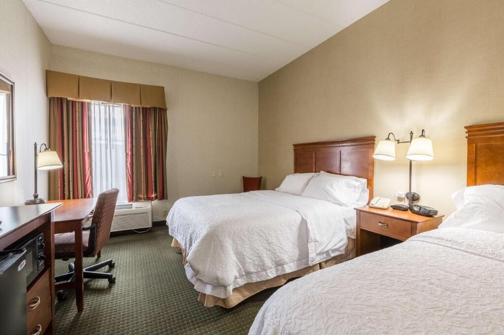 A hotel room with two beds, a desk, a chair, a lamp, and a window with curtains.