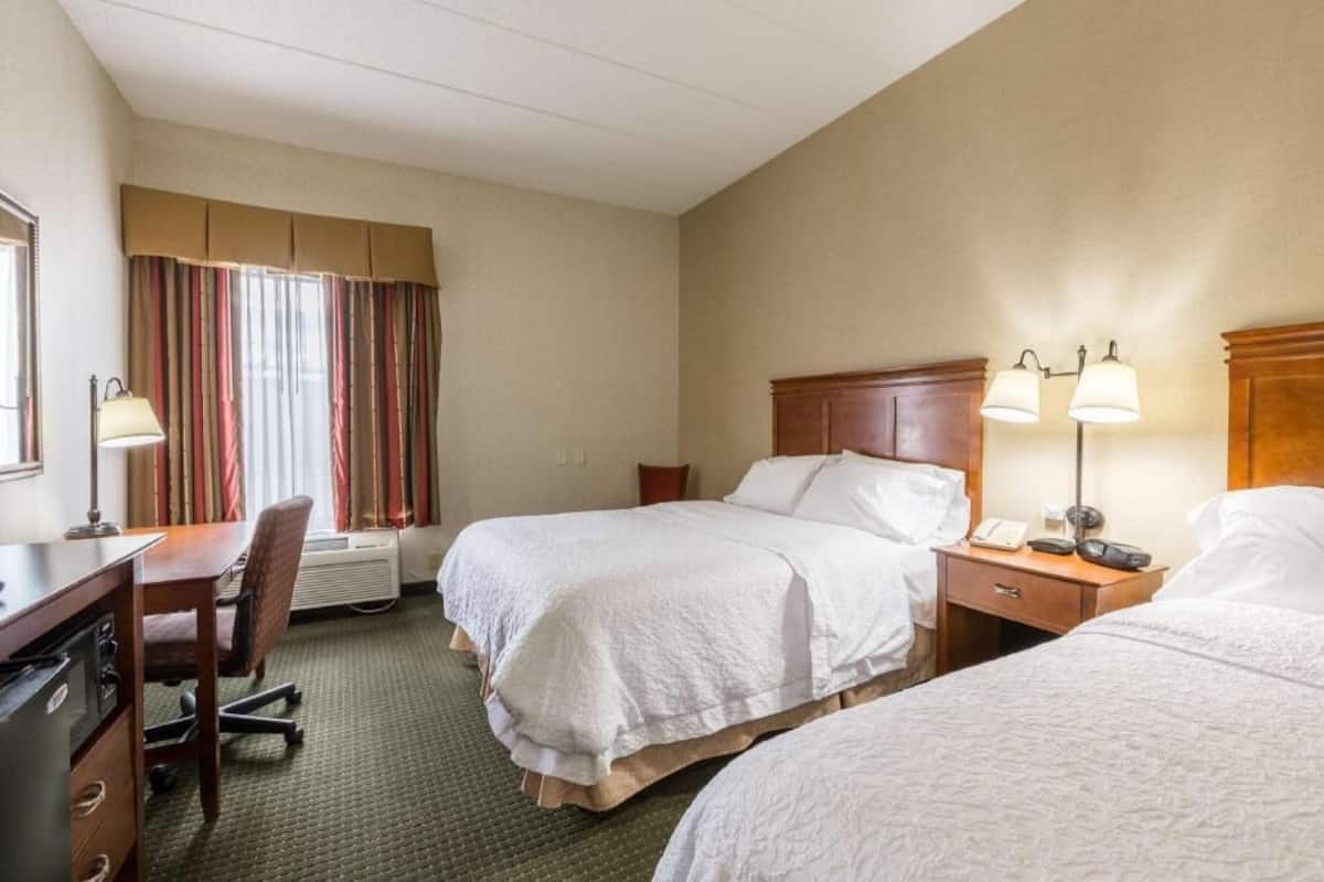 A hotel room with two beds, a desk, a chair, a lamp, and a window with curtains.