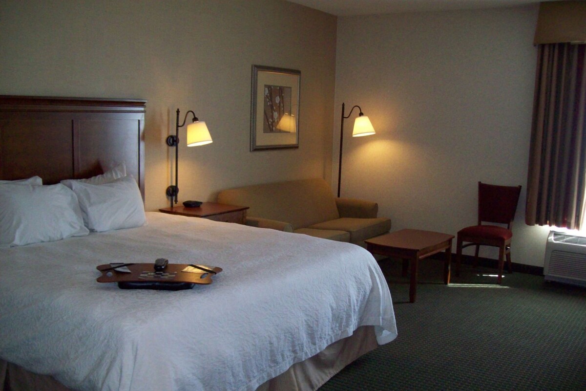 Habitación de hotel con una cama grande, un pequeño sofá, un escritorio y dos sillas. Hay dos lámparas de pared y un cuadro en la pared.