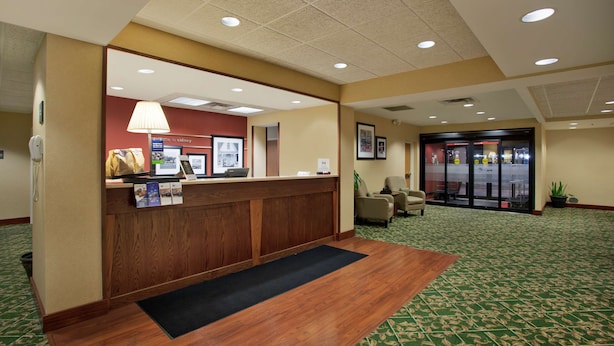 A hotel reception area with a wooden reception desk, a black mat, a green patterned carpet, and a glass door entrance. - Hampton Inn Sidney (Sidney)