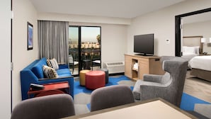 A hotel room with a blue sofa, a TV, a desk, a chair, a bed, and a view of the city.