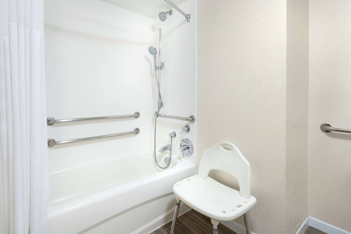 A bathroom with a white bathtub, grab bars, a showerhead, and a white chair.