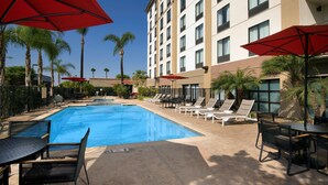 A hotel pool area with a rectangular pool, lounge chairs, and red umbrellas.